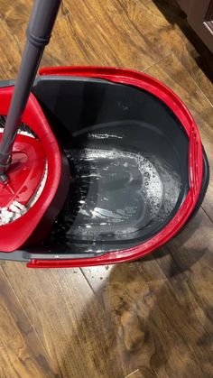 a red bucket filled with water on top of a wooden floor next to a black mop