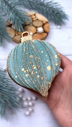 a hand holding a blue ornament with gold glitters on it next to some pine branches