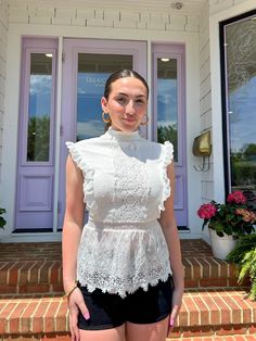 a woman standing in front of a door wearing black shorts and a white top with ruffles