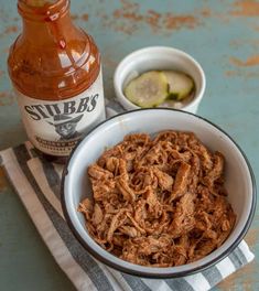 a white bowl filled with pulled pork next to a bottle of hot sauce and cucumber