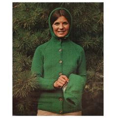 a woman wearing a green knitted sweater and hoodie standing in front of a pine tree