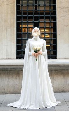 a woman in a white wedding dress and veil holding a bouquet stands on the sidewalk