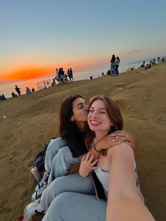 two women taking a selfie on the beach at sunset with people in the background