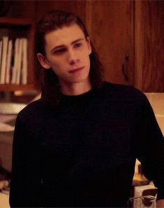 a man with long hair sitting at a table in front of a book shelf and looking off to the side