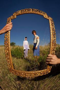 two people are standing in a field with a gold framed mirror on the ground and one person is taking a photo