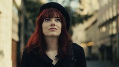 a woman with red hair wearing a black hat and looking off into the distance on a city street