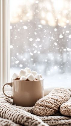 a cup of hot chocolate with marshmallows sitting on a blanket next to a window