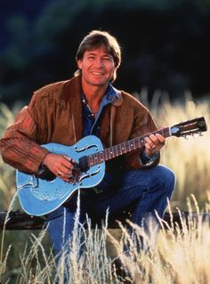 a man holding a blue guitar in his hands and smiling at the camera with tall grass behind him