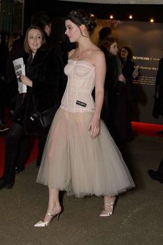 a woman in a dress standing on a red carpet next to other people and looking at something