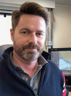 a man with a goatee is sitting in front of a computer desk and looking at the camera