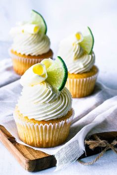 three cupcakes with white frosting and lime slices on top sitting on a wooden board