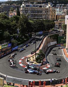 a group of cars driving around a track with buildings in the backgrouund