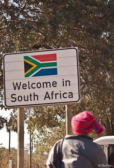 a sign that says welcome to south africa with a person walking by it and trees in the background