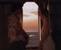 a man and woman standing next to each other in front of an open window at sunset