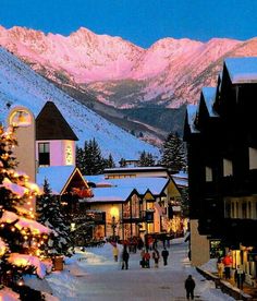 people are walking down the street in front of buildings with lights on them and snow covered mountains in the background