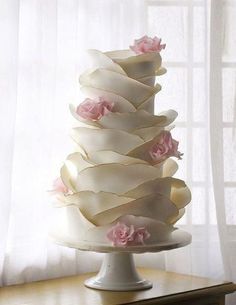 a three tiered white cake with pink flowers on the top and bottom, sitting on a table in front of a window