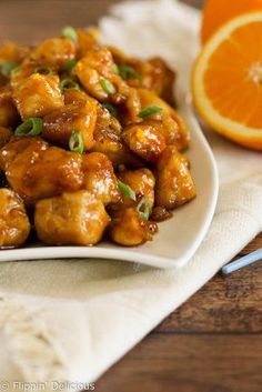 a white plate topped with orange chicken next to an orange wedge and fork on top of a wooden table