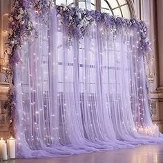 a room with sheer curtains, candles and flowers on the floor in front of it