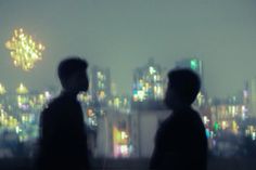 two people standing in front of a cityscape at night with fireworks coming from the sky