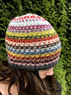 a woman wearing a multicolored knitted hat in front of green bushes and shrubbery