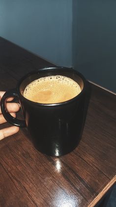 a cup of coffee sitting on top of a wooden table next to a person's hand