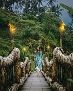 a woman walking across a wooden bridge surrounded by lush green hills and palm trees in the background