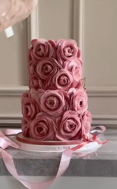 a three tiered cake with pink roses on top and ribbon around the edges, sitting on a table