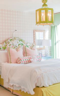 a white bed with pink and green pillows in a bedroom next to a lamp hanging from the ceiling