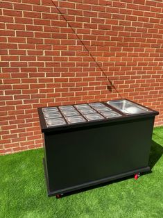 a black cooler sitting in the grass next to a brick wall with windows on it