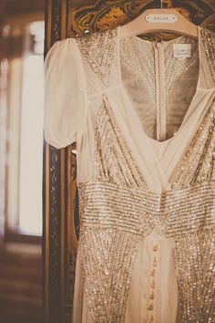 a white dress with gold sequins hanging on a wooden hanger in front of a mirror