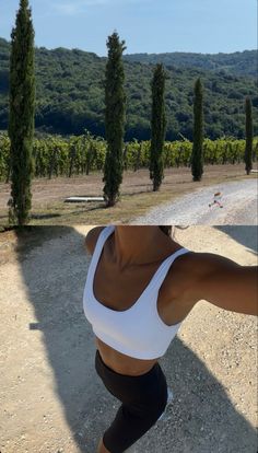 a woman in white shirt and black shorts standing on dirt road with trees behind her