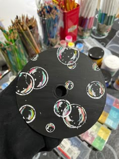 a person holding up a black circular object with circles painted on it and paintbrushes in the background