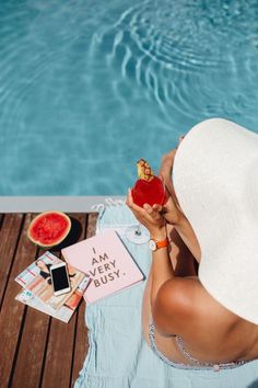 a woman in a white hat is sitting by the pool with her phone and watermelon