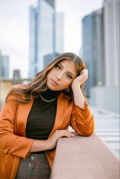 a beautiful young woman leaning on a wall in front of some tall buildings with her hand on her head