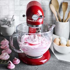 a red kitchen aid mixer with pink icing in the bowl next to eggs and utensils