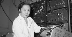 a woman in white lab coat standing next to an electronic equipment rack with wires on it