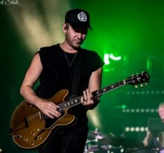 a man playing an electric guitar in front of a green stage light with other musicians behind him