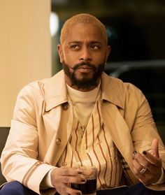 a man sitting down with a drink in his hand and looking at the camera while wearing a trench coat