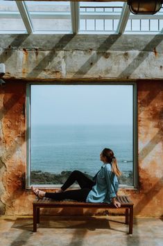 a woman sitting on top of a wooden bench next to a window near the ocean