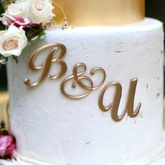 a white and gold wedding cake with flowers on the top is decorated in gold lettering