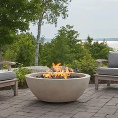 an outdoor fire pit sitting on top of a brick patio next to chairs and trees