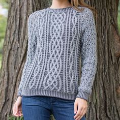 a woman standing in front of a tree wearing a gray knitted sweater and jeans