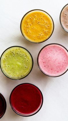 six different colored smoothies in small cups on a white surface, with the lids slightly open