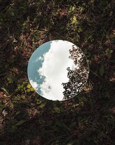 a round mirror sitting in the grass with a sky and clouds reflection on it's side