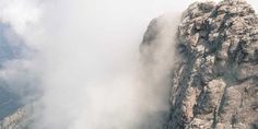 a person standing on top of a mountain next to a tall rock formation with clouds coming out of it