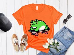 "Dive into the world of whimsy with our unique unisex t-shirt featuring a cute cartoon frog in a minimalist style, blissfully lying on a skateboard. This shirt blends playful charm with a sleek, modern design, making it perfect for anyone who loves unique and artistic clothing. Crafted for comfort and style, it's ideal for casual outings, skateboarding adventures, or simply as a trendy addition to your wardrobe. The shirt's design is not only eye-catching but also a conversation starter, appeali Frog Skateboard, Artistic Clothing, Cartoon Frog, Artist Outfit, Inspired Fashion, Minimalist Style, Family Shirts, Unisex Design, Minimalist Fashion