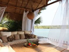 a living area with couches, pillows and curtains on the roof over looking water