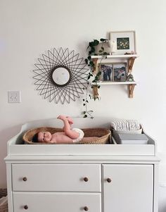 a baby laying in a basket on top of a dresser next to a wall mounted clock