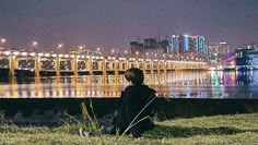 a person sitting on the grass looking out over water at night with lights in the background