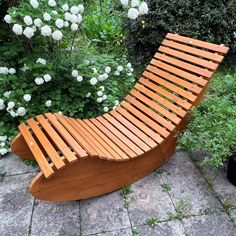 a wooden bench sitting on top of a sidewalk next to bushes and flowers in the background